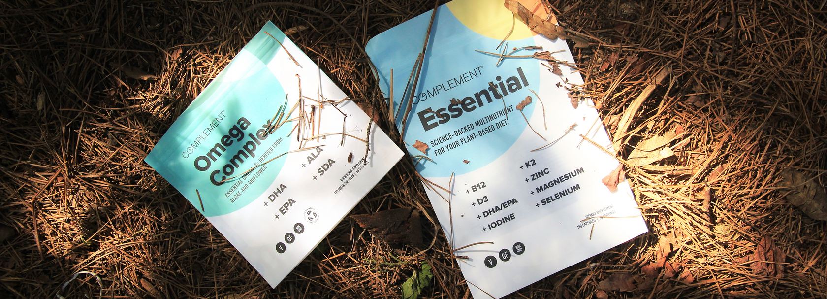 Two supplement packets labeled Omega Complex and Essential lie on the forest floor among pine needles and leaves, illuminated by dappled sunlight.